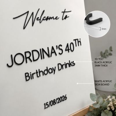 Personalised white acrylic birthday welcome sign with raised black lettering displayed on a wooden easel in an elegant event setting.